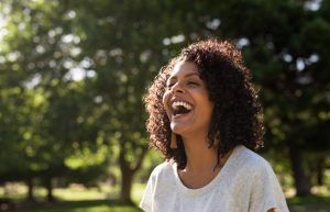 Woman with curly hair laughing outdoors, surrounded by trees.