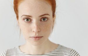 A young woman with red hair, green eyes, and freckles, wearing a white and black striped shirt, looks directly at the camera against a plain light background.