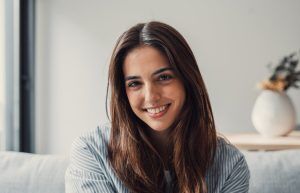 A young woman with long brown hair sits indoors, smiling at the camera. There is a white vase with dried flowers in the blurred background.