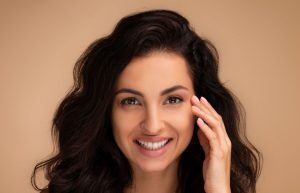 A woman with long, wavy dark hair smiles at the camera, her hand gently touching her face. She has clear skin and is photographed against a soft beige background.