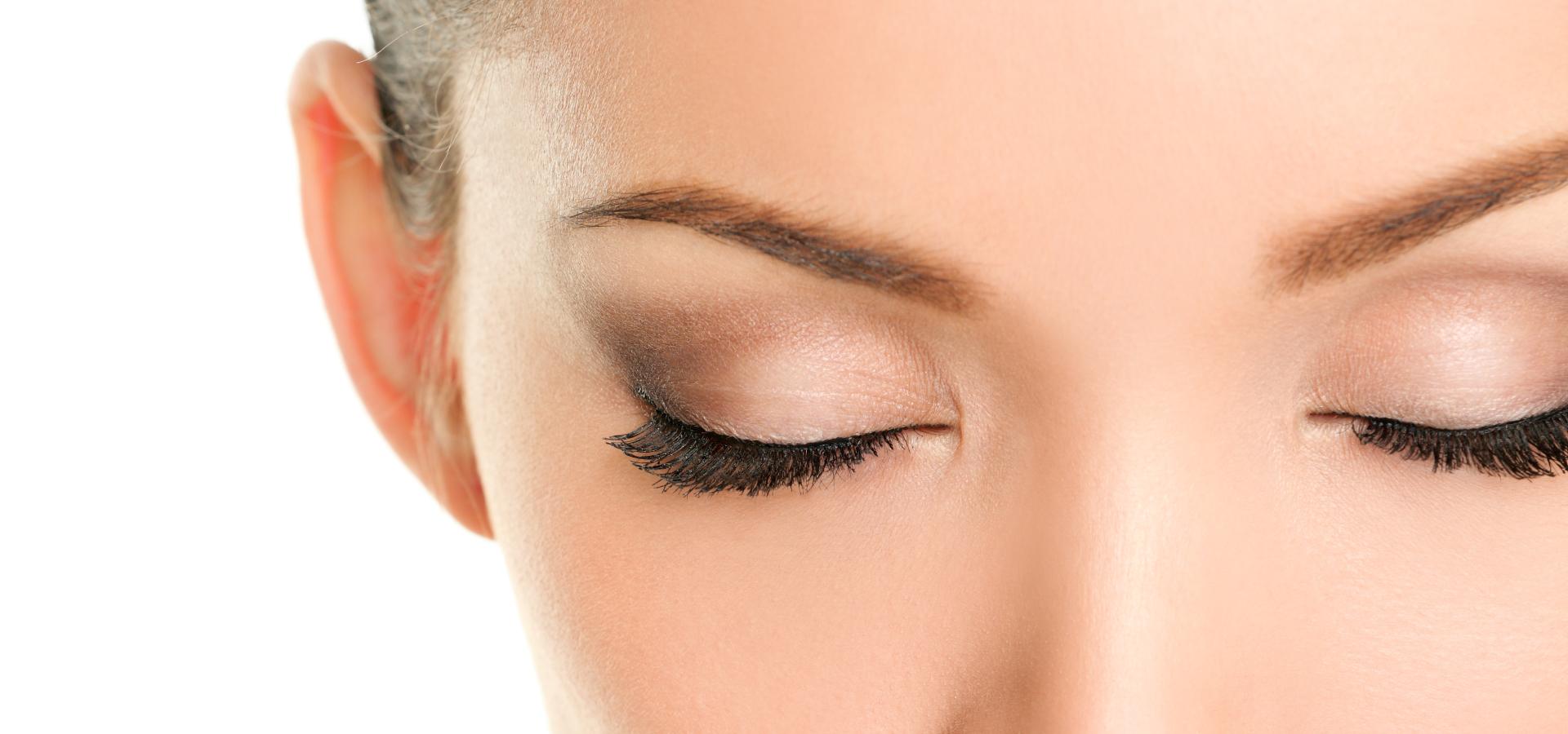 Close-up of a womans face with eyes closed, showing long black eyelashes and well-groomed eyebrows. The skin is smooth and makeup is applied to the eyelids. The background is plain white.