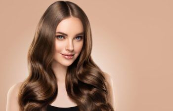 A woman with long, shiny, wavy brown hair and blue eyes smiles softly against a light beige background. She is wearing a black top.