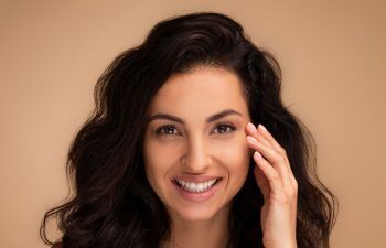 A woman with long, wavy dark hair smiles at the camera, her hand gently touching her face. She has clear skin and is photographed against a soft beige background.