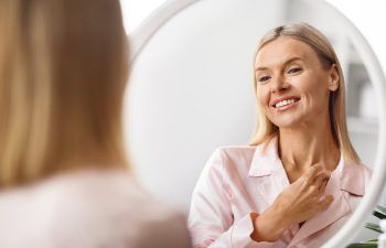 A woman in pink pajamas smiles as she looks at herself in a round mirror, touching her neck gently with one hand.