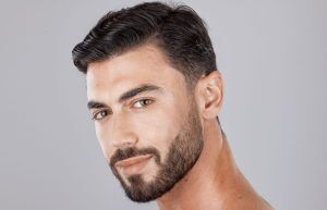 A man with short dark hair and a neatly trimmed beard looks at the camera with a slight smile, against a plain gray background.