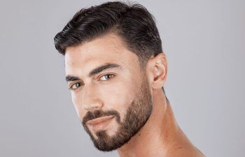 A man with short dark hair and a neatly trimmed beard looks at the camera with a slight smile, against a plain gray background.